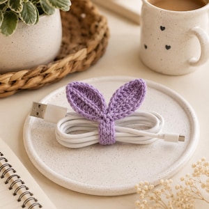 Könnte beinhalten: Ein weißes USB-Ladekabel, das aufgewickelt und mit einem lavendelfarbenen Häkel-Kabelorganizer befestigt ist, der zwei aufrechte, blattförmige Hasenohren aufweist. Das Kabel hat einen Standard-USB-A-Anschluss und einen kleineren Anschluss.