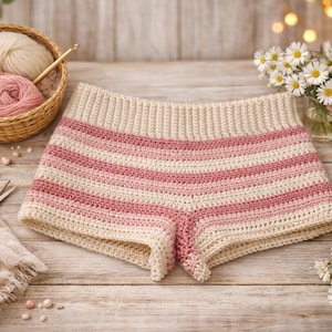 May include: Crochet shorts in cream and pink stripes. The shorts are displayed on a wooden surface, with a basket of yarn, scissors, and daisies. The shorts have a ribbed waistband and a striped pattern.