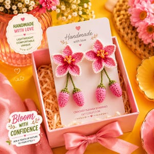 Könnte beinhalten: Ein Paar gehäkelte Blumenohrringe in Pink mit weißen Rändern und gelben Zentren, befestigt an einer weißen Karte mit der Aufschrift Crochet Flower Earrings Lily Collection. Die Ohrringe haben hängende pinke Knospen an grünen Stielen.