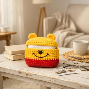 Könnte beinhalten: Eine gelb, rot und schwarz gehäkelte Hülle mit einem Bärengesicht-Design. Die Hülle befindet sich auf einem weißen Holztisch mit einer Tasse, einer Brille und einem Untersetzer. Die Hülle hat ein lächelndes Gesicht und Bärenohren.