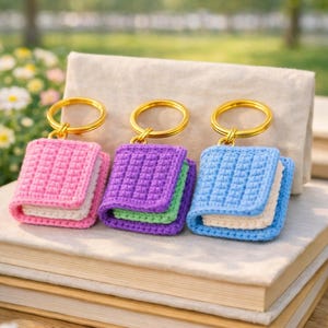 Könnte beinhalten: Drei gehäkelte, buchförmige Schlüsselanhänger in Rosa, Lila und Blau, jeweils mit einem goldenen Schlüsselring. Die Schlüsselanhänger liegen auf einem Stapel Bücher mit verschwommenem Hintergrund aus Grün.