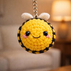 May include: A crocheted bee keyring with a yellow body, black stripes, and white wings. The bee has a smiling face with black eyes and pink cheeks. The keyring is attached to a silver metal ring and chain.