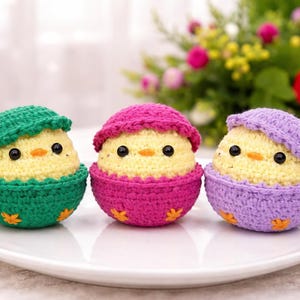 May include: Three handmade crochet chicks in eggshell-shaped containers. Each chick is yellow with black eyes and an orange beak, wearing a matching hat. The containers are green, pink, and purple, with orange star accents. The chicks sit on a white plate.