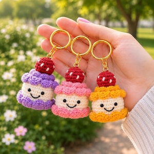 Könnte beinhalten: Drei gehäkelte Schlüsselanhänger in Form lächelnder Cupcakes mit roten Erdbeerspitzen. Jeder hat eine cremefarbene Basis, eine strukturierte Glasurschicht in Lila, Rosa oder Gelb und einen goldfarbenen Metallring zur Befestigung an Schlüsseln.