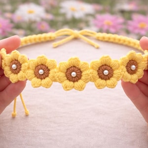 Könnte beinhalten: Ein handgefertigtes, gelbes Häkelstirnband mit fünf Sonnenblumenmotiven mit braunen Zentren und weißen Perlenakzenten. Das Stirnband wird von zwei Händen gehalten, mit einem verschwommenen floralen Hintergrund.