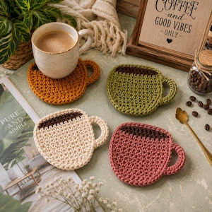 May include: Four handmade crochet coasters shaped like coffee mugs in mustard yellow, olive green, cream, and dusty rose. Each coaster features a dark brown top section representing coffee and a small handle on the right side, crafted with yarn.