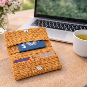Könnte beinhalten: Eine senffarbene gehäkelte Geldbörse mit Druckknopfverschluss, die einen blauen Reisepass und zwei Karten enthält. Ein Laptop, eine weiße Tasse mit Tee und eine Vase mit rosa Rosen befinden sich im Hintergrund, alles auf einer Holzoberfläche.