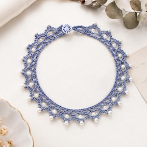 May include: A delicate, blue crocheted necklace with a scalloped edge and small, white pearl accents. The necklace is circular and fastened with a small, decorative button. The design is intricate and elegant, suitable for special occasions.