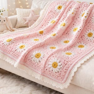 Può includere: Una coperta all'uncinetto rosa e bianca con grandi motivi a margherita. La coperta ha un bordo smerlato ed è drappeggiata su un divano bianco. Un vaso di fiori, un coniglietto di peluche e un giocattolo di legno sono visibili.