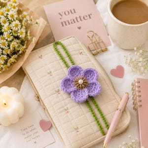 Könnte beinhalten: Ein cremefarbenes, gestepptes Stoffetui mit kleinem Blumenmuster und einem grünen Häkelband, das mit einer violetten Häkelblume verziert ist. Die Blume hat eine braune Mitte und eine kleine weiße Perle, die als Lesezeichen dient.