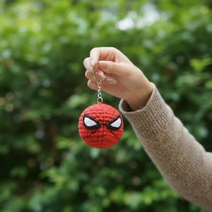 Op de afbeelding: Een rood en zwart gehaakte Spider-Man sleutelhanger. De ronde sleutelhanger heeft witte ogen en een webpatroon. De sleutelhanger wordt omhoog gehouden tegen een vage groene achtergrond.