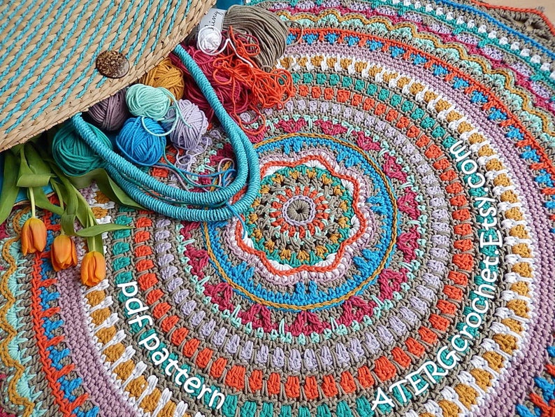 May include: A colorful crochet mandala rug with a variety of colors, including orange, red, pink, blue, green, and yellow. The rug is made with a variety of stitches and patterns, and it has a unique and intricate design. The rug is surrounded by yarn balls and a wicker bag.