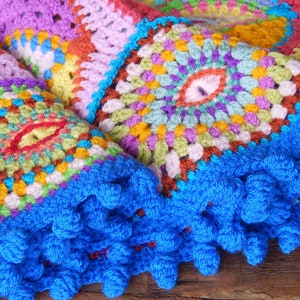 May include: A colorful crocheted blanket with a blue fringe border. The blanket is made up of squares with different patterns and colors, including red, orange, yellow, green, blue, purple, and pink.
