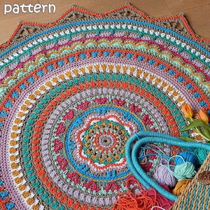 May include: A colorful crochet mandala rug with a starburst pattern in shades of orange, red, green, blue, and brown. The rug is made with a variety of stitches and textures, creating a unique and eye-catching design. The rug is laid out on a wooden floor.