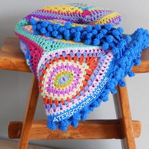 May include: A colorful crocheted blanket with a blue pom-pom trim. The blanket is made with a variety of colors, including pink, purple, green, yellow, and orange. The blanket is folded and draped over a wooden stool.