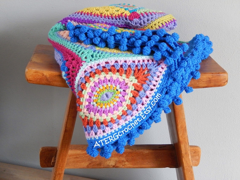 May include: A colorful crocheted blanket with a blue pom-pom trim. The blanket is made with a variety of colors, including pink, purple, green, yellow, and orange. The blanket is folded and draped over a wooden stool.