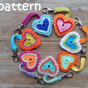 May include: Seven crocheted heart keychains in various colours, including pink, orange, blue, green, and white, with silver key rings and colourful ribbon loops. The keychains are arranged in a circle on a wooden surface. The text "pattern" is visible at the top of the image.
