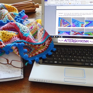May include: A colorful crocheted blanket with blue pom-poms lies on a table next to a laptop computer with a crochet pattern displayed on the screen. The text on the screen reads "~ATERGcrochet~".