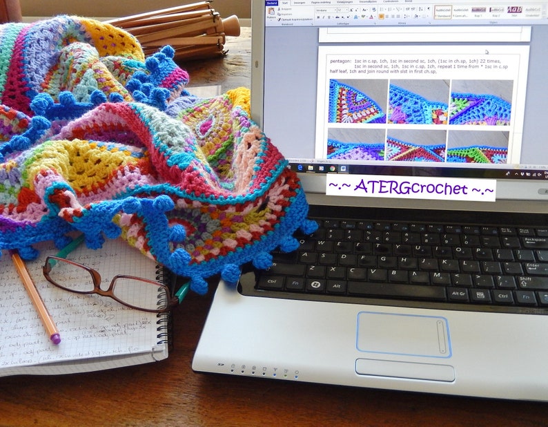 May include: A colorful crocheted blanket with blue pom-poms lies on a table next to a laptop computer with a crochet pattern displayed on the screen. The text on the screen reads "~ATERGcrochet~".