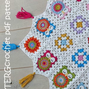 Peut inclure: Une couverture en crochet blanche avec des motifs de hiboux colorés et des glands. La couverture est faite de carrés de grand-mère et présente une variété de couleurs, notamment le rose, le bleu, le vert, le jaune et l'orange.