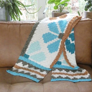 May include: A crocheted throw blanket with a geometric pattern in shades of blue, white, and brown. The blanket is folded and draped over a brown leather couch.
