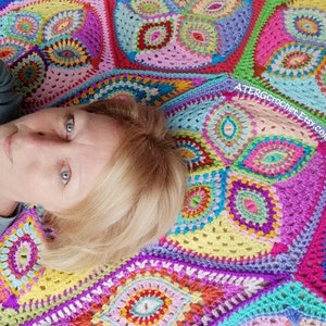 May include: A woman lies on a colorful crocheted blanket with a multi-colored mandala pattern. The blanket is made of yarn in shades of pink, purple, blue, green, yellow, and orange. The blanket has a website address, "ATERGcrochet.Etsy.com", printed on it.
