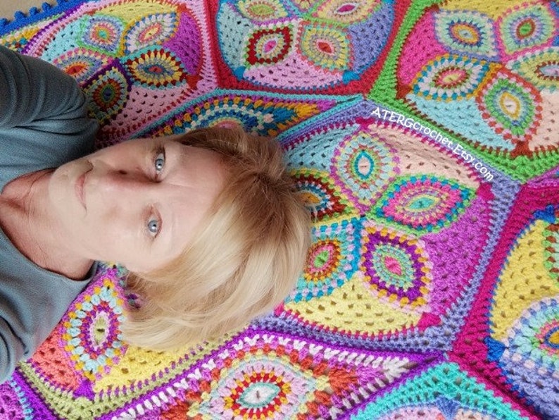 May include: A woman lies on a colorful crocheted blanket with a multi-colored mandala pattern. The blanket is made of yarn in shades of pink, purple, blue, green, yellow, and orange. The blanket has a website address, "ATERGcrochet.Etsy.com", printed on it.