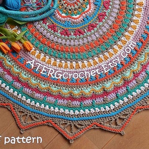 May include: A colorful crochet rug with a mandala design. The rug is made with a variety of colors, including orange, red, green, blue, and brown. The rug has a fringe border and is laid out on a wooden floor. The text "ATERGcrochet Etsy.com" is visible in the center of the rug. The text "pdf pattern" is visible at the bottom of the image.