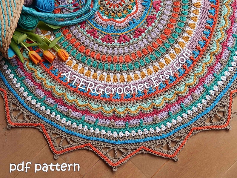 May include: A colorful crochet rug with a mandala design. The rug is made with a variety of colors, including orange, red, green, blue, and brown. The rug has a fringe border and is laid out on a wooden floor. The text "ATERGcrochet Etsy.com" is visible in the center of the rug. The text "pdf pattern" is visible at the bottom of the image.