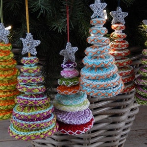 May include: Crocheted Christmas tree ornaments in various colors, including orange, green, blue, and purple. Each tree is topped with a silver glitter star and has a unique stitched design. The ornaments are hanging in front of a wicker basket.