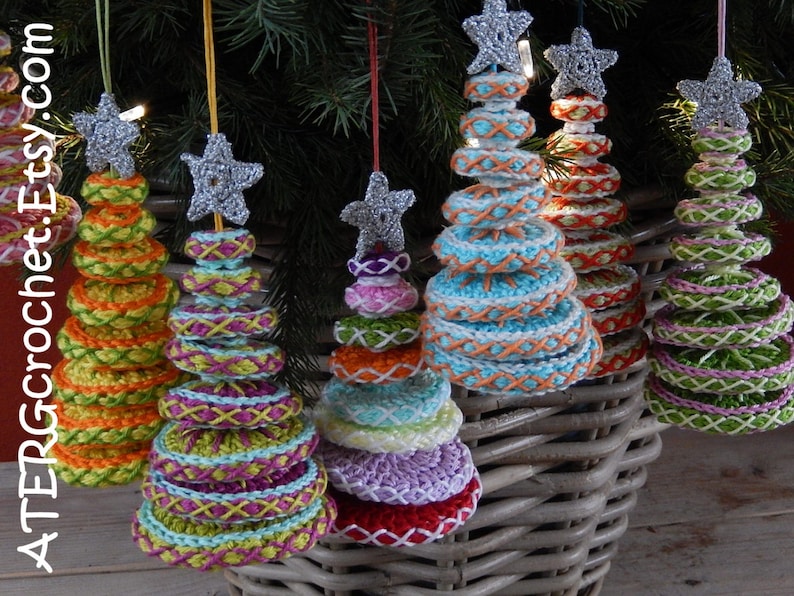 May include: Crocheted Christmas tree ornaments in various colors, including orange, green, blue, and purple. Each tree is topped with a silver glitter star and has a unique stitched design. The ornaments are hanging in front of a wicker basket.
