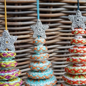 May include: Three handmade crochet Christmas tree ornaments. Each ornament features a silver star on top, with colorful stacked rings in shades of teal, pink, orange, and white. The ornaments are suspended by colorful strings.