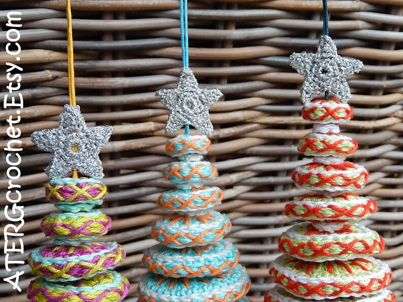 May include: Three handmade crochet Christmas tree ornaments. Each ornament features a silver star on top, with colorful stacked rings in shades of teal, pink, orange, and white. The ornaments are suspended by colorful strings.