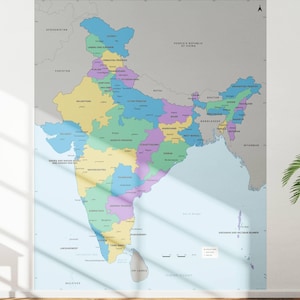 Puede incluir: Un mapa grande y colorido de la India, exhibido en una pared blanca. El mapa está dividido en varios estados, cada uno distinguido por un color diferente. Una pequeña planta en maceta se encuentra en un taburete de madera a la derecha del mapa.