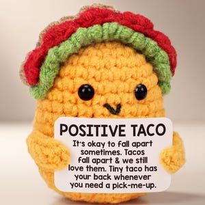 May include: A yellow crocheted taco plush toy with black bead eyes and a stitched mouth. It wears a brown, green, and red crocheted topping. It holds a white sign that reads: POSITIVE TACO. It's okay to fall apart sometimes. Tacos fall apart & we still love them. Tiny taco has your back whenever you need a pick-me-up.