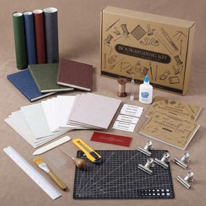 May include: A complete bookbinding kit with various tools and materials. Includes a cardboard box with the text "BOOKBINDING KIT", notebooks, paper, glue, a cutting mat, and other crafting supplies. The kit also contains a ruler, brush, and metal clips.