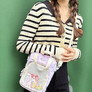May include: A light purple crossbody bag with a mesh front panel featuring a cartoon graphic. The bag has a top handle and a white shoulder strap. The person is wearing a black and beige striped cardigan.