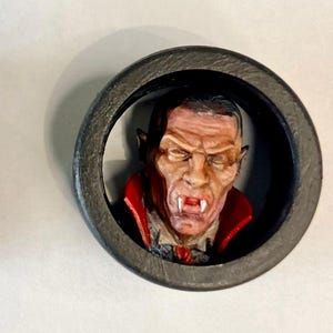 May include: Two black, circular decorative pieces each feature a detailed, miniature bust of a vampire. The vampire has pale skin, red accents, and a stern expression. The busts are set against a dark background.