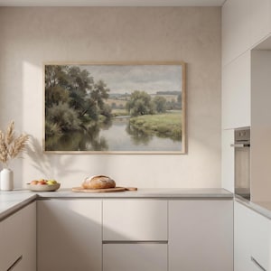 Könnte beinhalten: Kücheninterieur mit einem gerahmten Landschaftsbild über einer Arbeitsplatte. Eine Vase mit getrockneten Pflanzen, eine Schale mit Obst und ein Brotlaib auf einem Brett befinden sich auf der Theke. Die Küche hat weiße Schränke und ein Fenster.
