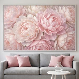 May include: A large rectangular wall art print featuring a dense arrangement of soft pink and cream-colored peony flowers in full bloom. The artwork is encased in a thin white frame and displayed above a gray sofa with matching pink throw pillows.