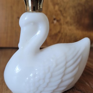May include: A white swan figurine with a gold-colored crown on its head. The swan has a smooth, glossy finish and detailed feather patterns on its wings. The figurine is likely made of ceramic or glass and is a decorative item.