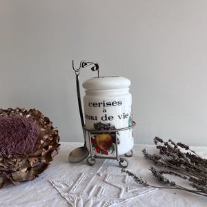 May include: A white ceramic jar with a lid, featuring the text "cerises à eau de vie" and fruit illustrations. It sits in a metal stand with a spoon. A dried flower and lavender sprigs are also present.