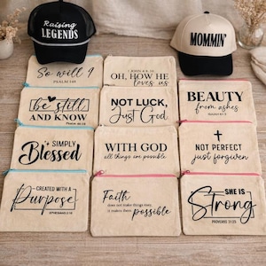May include: A collection of canvas zipper pouches and baseball caps. The pouches feature inspirational quotes and Bible verses in black script. The caps have the words "Raising Legends" and "Mommin" embroidered on them. The pouches have colored zippers.