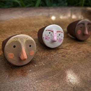 May include: Three rings featuring hand-painted faces on circular brown and white bases. Each ring displays distinct facial features including painted eyes, noses, and rosy cheeks in shades of pink, orange, black, and brown on a dark brown band.