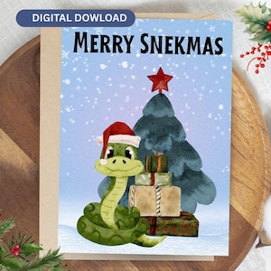 Può includere: Un biglietto d'auguri con un serpente verde che indossa un cappello da Babbo Natale rosso e bianco, accanto a una pila di regali incartati e un albero di Natale verde scuro con una stella rossa. Il testo MERRY SNEKMAS è stampato in nero in alto.