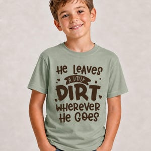 May include: A sage green short-sleeve crew neck t-shirt featuring dark brown text that reads He Leaves A Little Dirt Wherever He Goes, with the word Dirt in a larger, bold font and decorative splatter accents surrounding the lettering.
