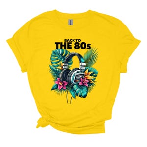 May include: A bright yellow t-shirt featuring a graphic print of black headphones with a cassette tape design, surrounded by colorful tropical leaves and pink flowers, with the black text BACK TO THE 80s printed above the headphones.