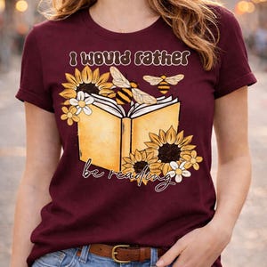 May include: Burgundy t-shirt with the text "I would rather be reading." The graphic features an open book with sunflowers, daisies, and bees. The shirt has short sleeves and a crew neck.