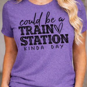 Könnte beinhalten: Ein lila T-Shirt mit dem schwarzen Aufdruck "could be a TRAIN STATION kinda day". Das Shirt hat einen Rundhalsausschnitt und kurze Ärmel. Der Text enthält ein Herzsymbol.