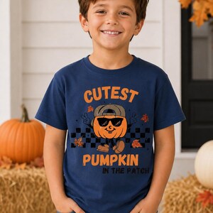 May include: A navy blue short-sleeve t-shirt featuring a graphic of an orange pumpkin wearing sunglasses and a baseball cap, making a peace sign. The text reads CUTEST PUMPKIN IN THE PATCH, accompanied by a black and white checkered pattern.
